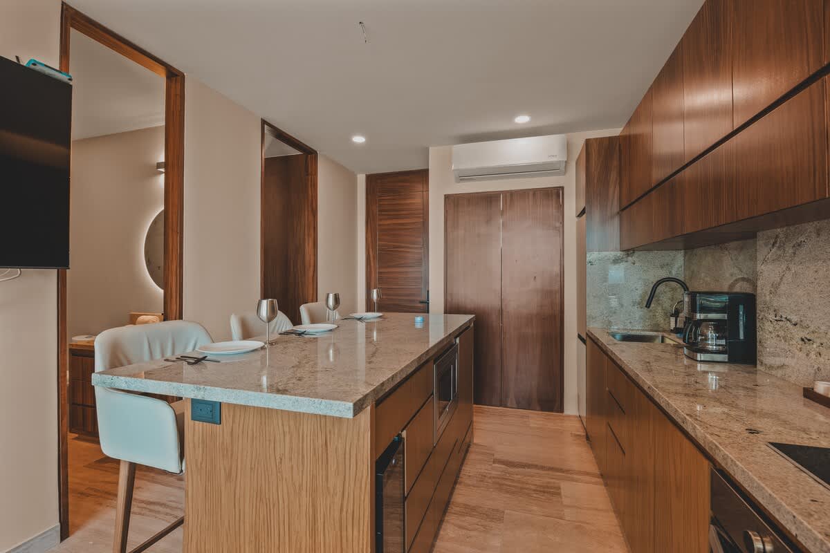 Modern kitchen with a large granite-topped island serving as a dining bar with two white stools, integrated microwave, and dark wood cabinetry, complemented by light-colored walls and flooring.