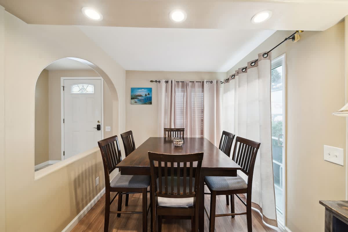 A view of the dining area shows a dark wood table with six chairs, set on wood-look flooring, with light-colored curtains on the far wall. An arched opening leads to the front door area, and recessed lighting brightens the room.