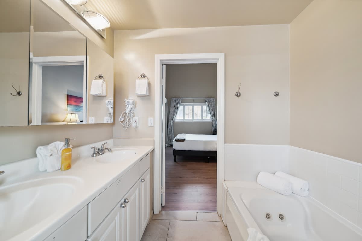 A bright bathroom with light beige walls and tile flooring, featuring a double vanity with white cabinets and two sinks. A large mirror with vanity lighting is above the sinks, and a jetted bathtub is partially visible on the right. An open doorway leads to a bedroom.