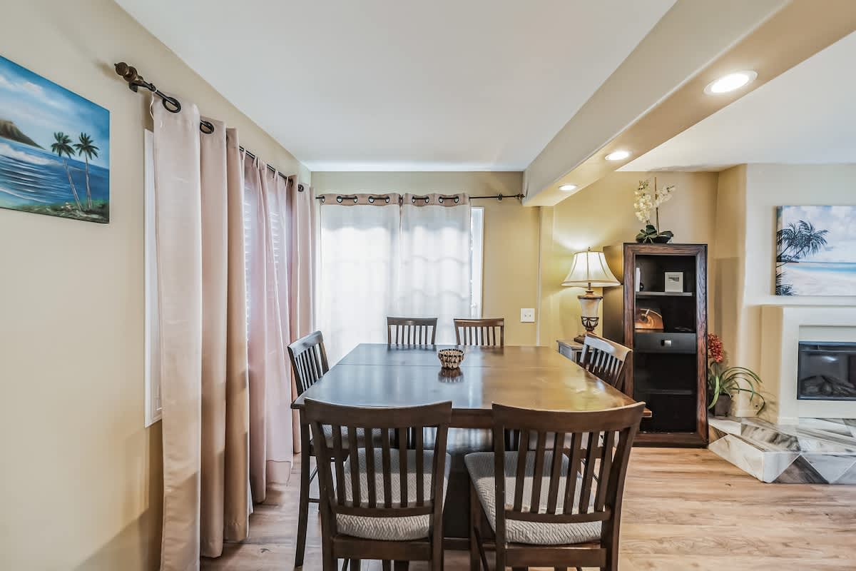 This dining room features a large dark wood table with six high-back chairs, positioned on wood-look flooring. Light-colored curtains frame a window, and a decorative artwork hangs on the wall, with recessed lighting illuminating the space.