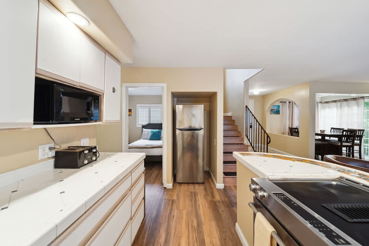 View of the kitchen showing a stainless steel refrigerator, a doorway leading to a bedroom, and a staircase in the background, all set against warm beige walls and hardwood flooring.