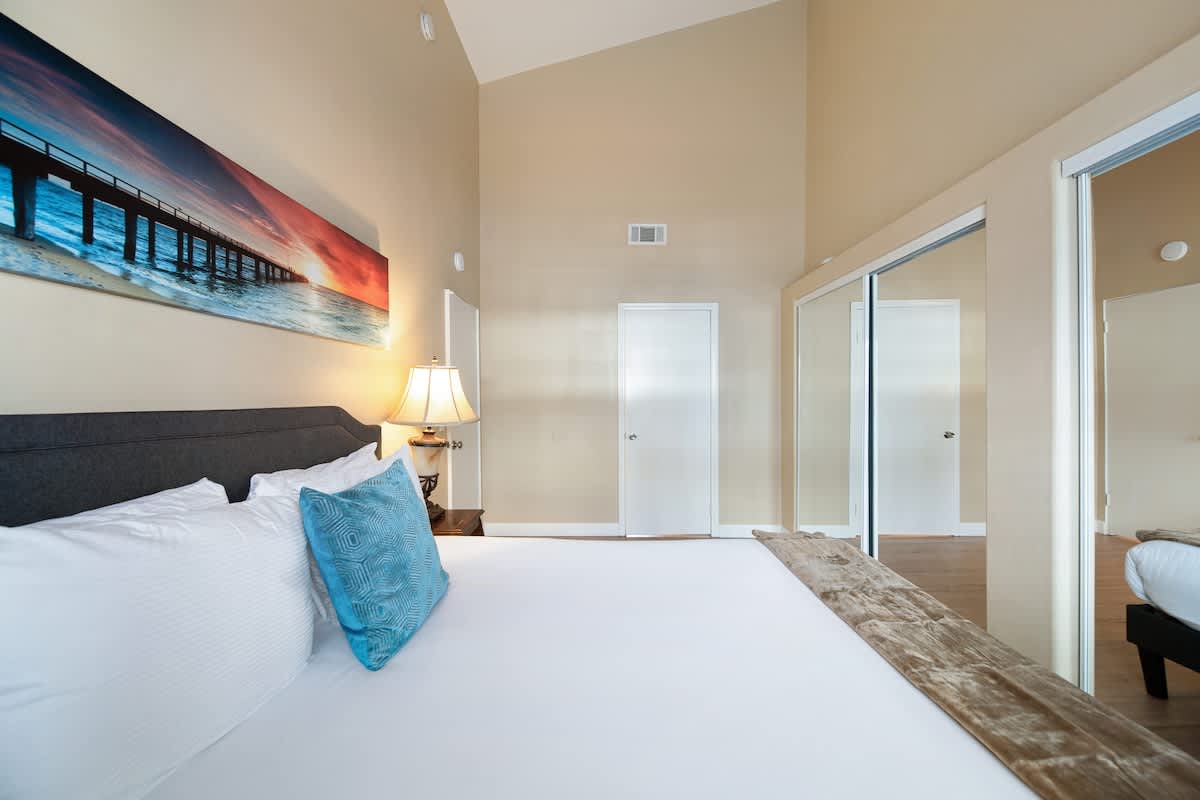 A bedroom with light beige walls and a vaulted ceiling, featuring a queen-sized bed with a dark gray headboard, white bedding, and a blue accent pillow. A large landscape artwork hangs above the bed, and a mirrored closet is visible on the right, reflecting the room.