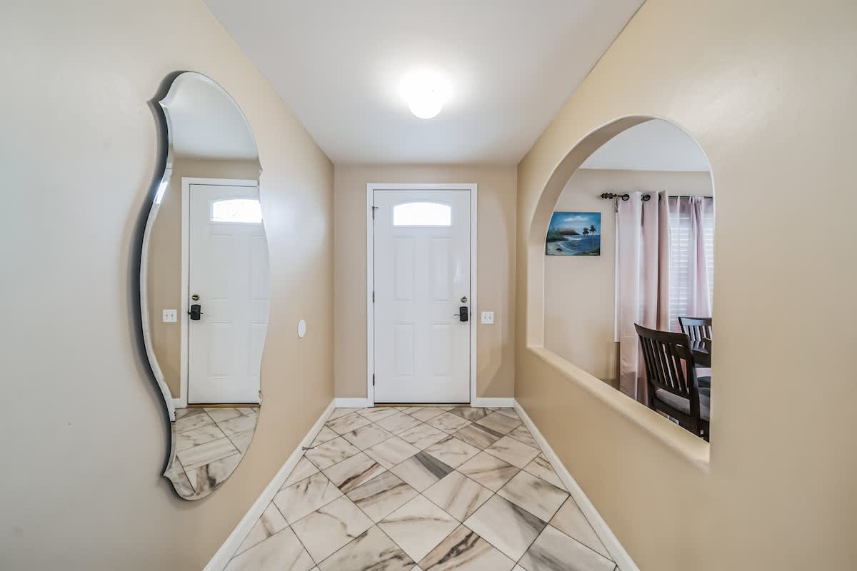 A welcoming entryway with light-colored marble-look tiled flooring laid in a diagonal pattern, a large decorative mirror reflecting the front door, and an arched opening leading to another room.