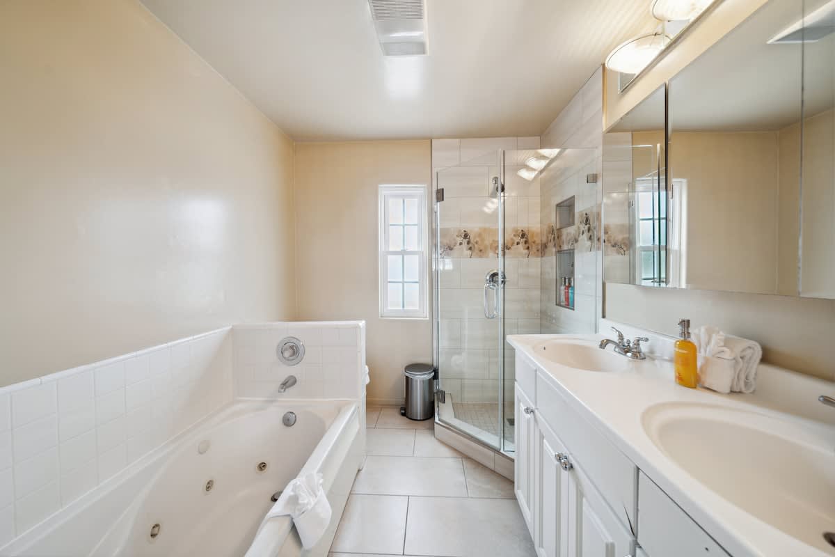 A spacious bathroom with light beige walls and light-colored tile flooring, featuring a large jetted bathtub on the left and a glass-enclosed shower on the right. A double vanity with white cabinets and two sinks is visible, along with a window above the tub.