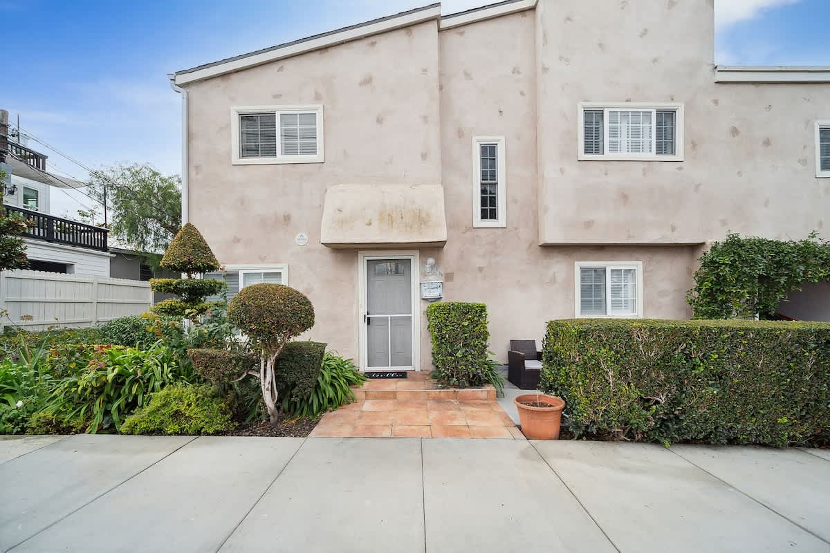The front exterior of this two-story house showcases a light-colored stucco finish with several windows and a central entrance shaded by an awning. Well-maintained green landscaping, including hedges and ornamental shrubs, borders the property, with a paved walkway leading to the door.