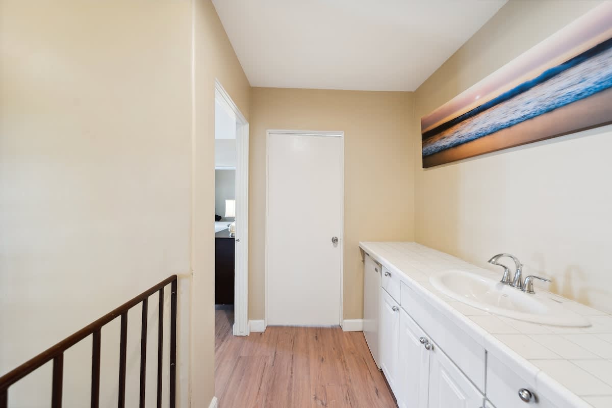 A hallway featuring a white vanity with a built-in sink and tiled countertop, a long horizontal artwork on the wall, and a doorway leading to another room.