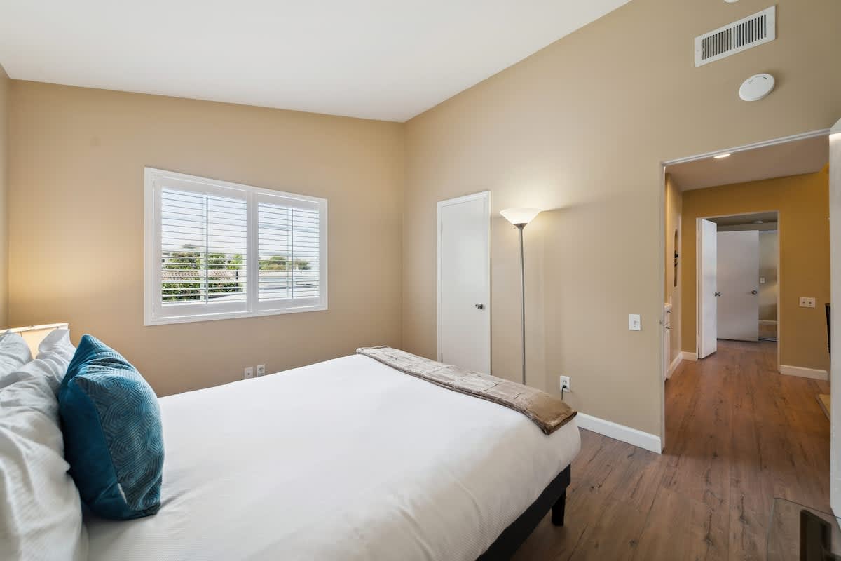 A cozy bedroom with light beige walls and dark wood laminate flooring, featuring a queen-sized bed with white bedding and a blue accent pillow. A window with white plantation shutters is visible, and a tall floor lamp stands near a white door.