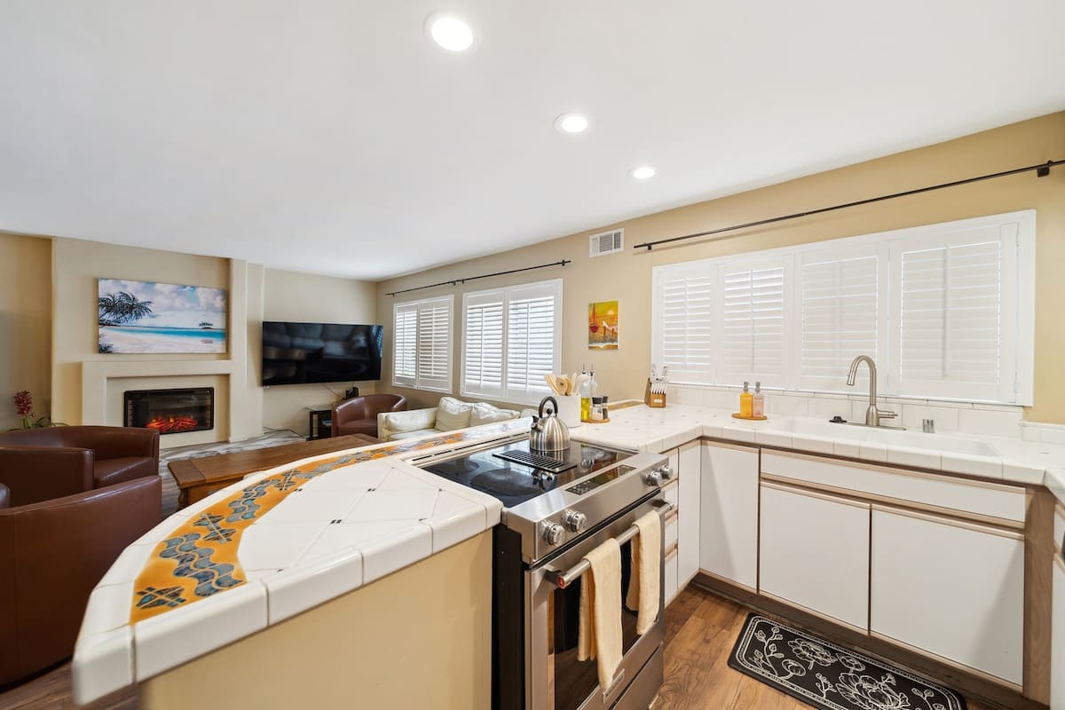 An open-concept kitchen with white tiled countertops and cabinets, a stainless steel range, and a decorative tiled breakfast bar, flowing into a living room with a fireplace and a large wall-mounted TV.