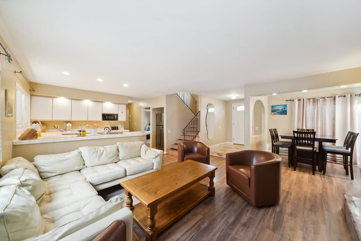 A wide view of the open-concept living area shows a white sectional sofa, a wooden coffee table, and two brown leather armchairs on dark wood-look flooring. In the background, a dining area with a dark wood table and chairs is visible, along with a partial view of the kitchen counter.