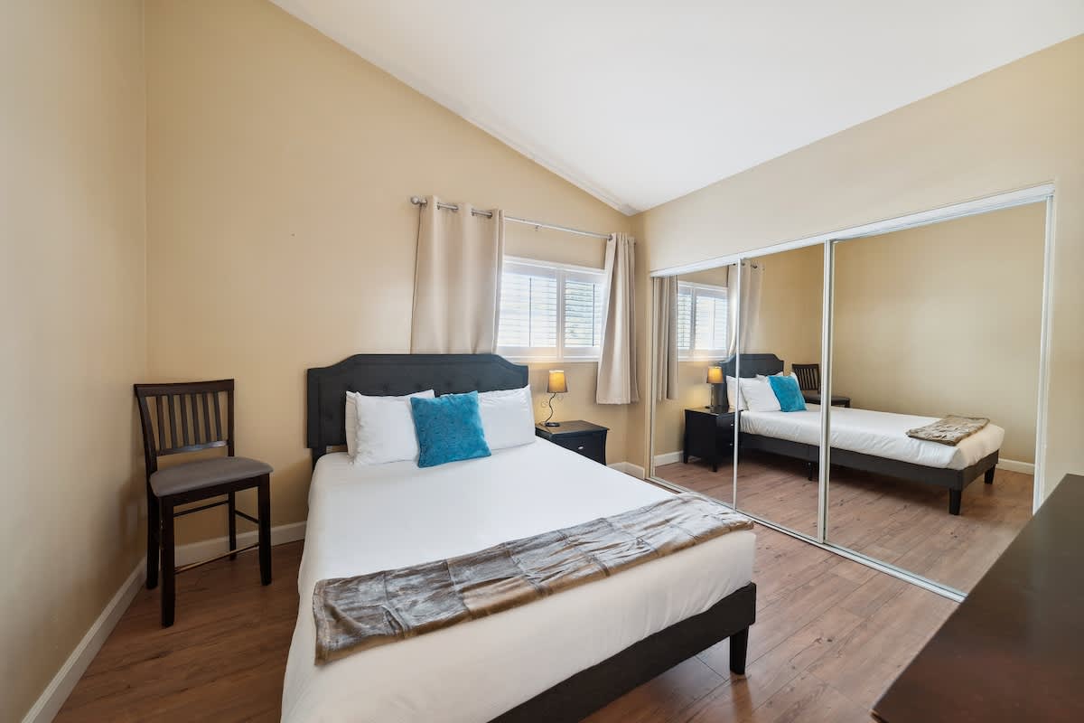 A bright bedroom with light beige walls, a vaulted ceiling, and dark wood laminate flooring. It features a queen-sized bed with a dark gray headboard, white bedding, a blue accent pillow, and a brown throw. A large mirrored closet spans one wall, reflecting the room.