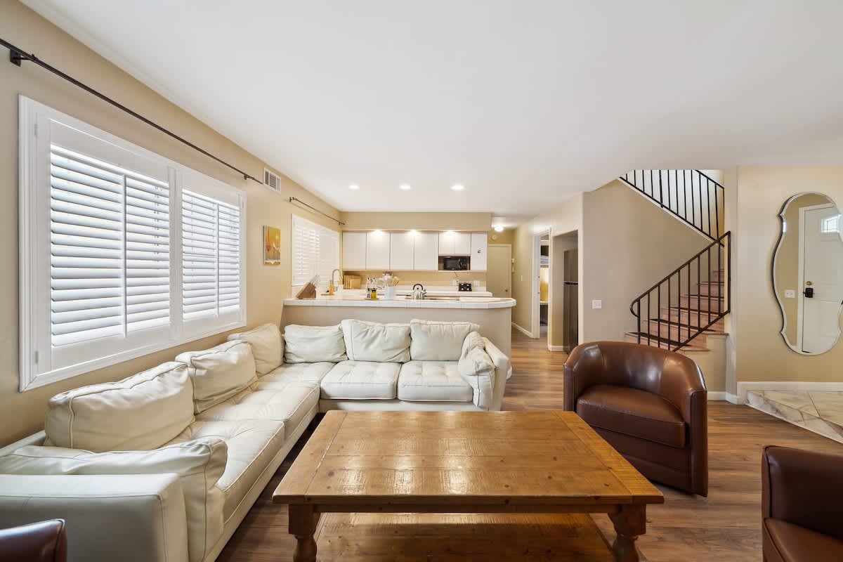 An open-concept living room with light beige walls and dark wood laminate flooring, featuring a large white sectional sofa with a brown leather armchair. A rustic wooden coffee table is centered in the room, which opens to a kitchen area with white cabinets and a breakfast bar.