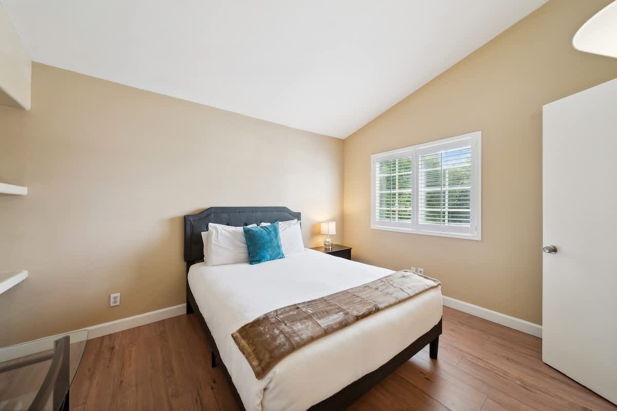 A bright bedroom with light beige walls, a vaulted ceiling, and dark wood laminate flooring. A queen-sized bed with a dark gray headboard, white bedding, a blue accent pillow, and a brown throw is centered. A window with white plantation shutters offers a view of greenery.