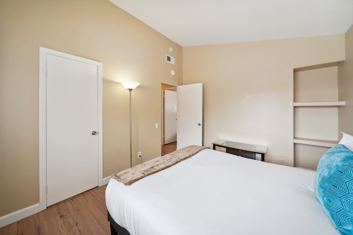 A bedroom with light beige walls and dark wood laminate flooring, showing the foot of a queen-sized bed with white bedding and a brown throw. A tall floor lamp stands between two white doors, and built-in shelves are recessed into the wall on the right.
