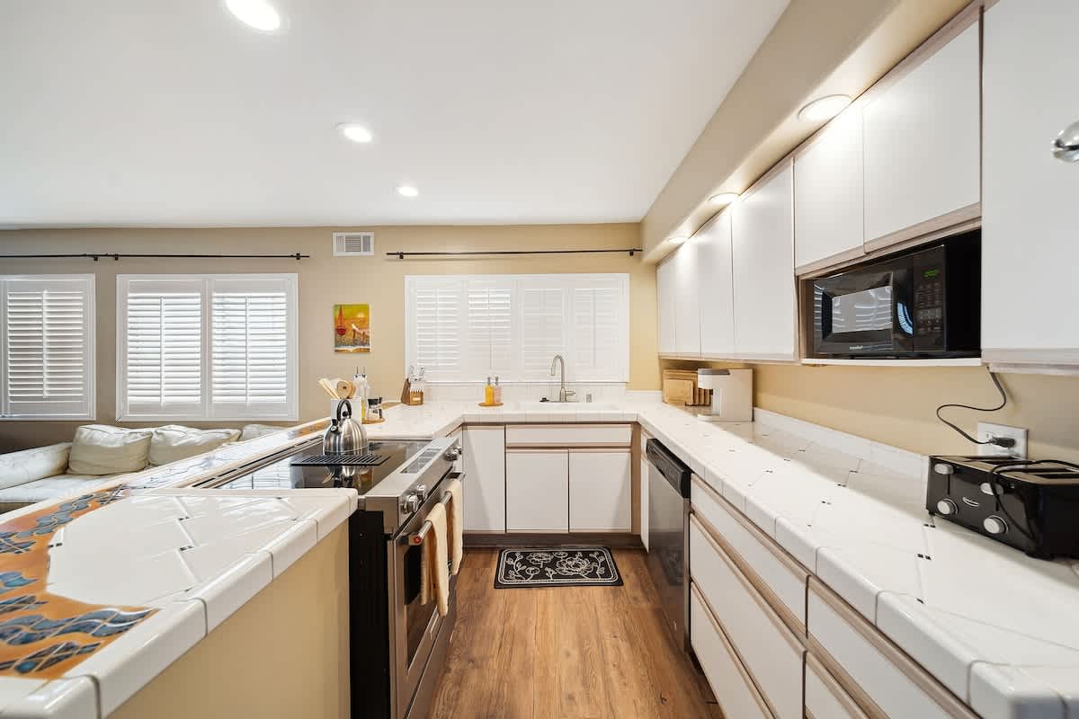 A bright kitchen featuring white tiled countertops, white upper and lower cabinets, and a stainless steel range with a built-in microwave above. The kitchen opens to a living area with white plantation shutters.