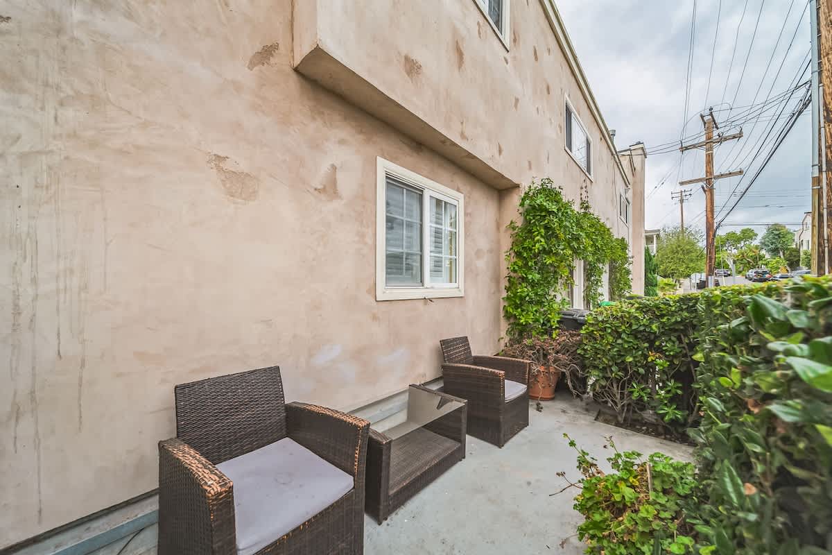 An outdoor patio area with concrete flooring, featuring two dark brown wicker armchairs with light cushions and a matching small table. The area is bordered by green hedges and the exterior wall of the building, with a window visible above.