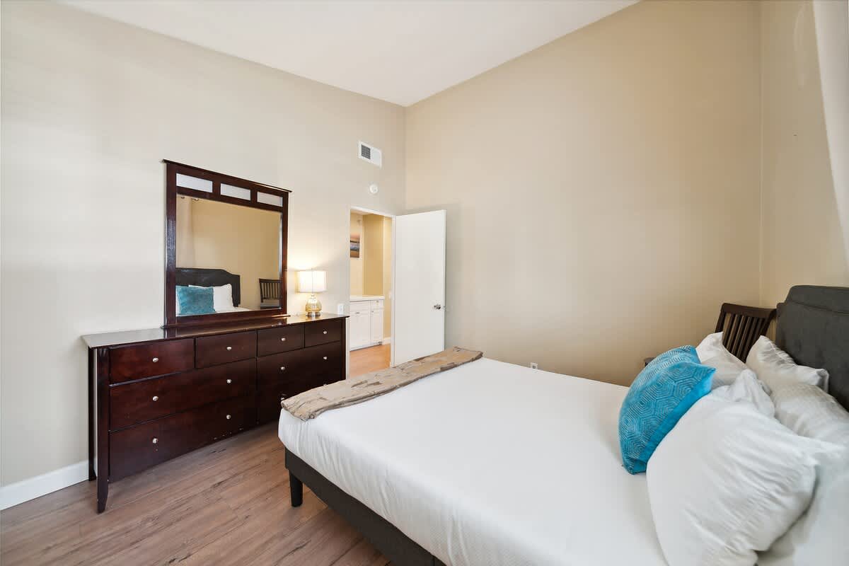 A bedroom featuring a dark wood dresser with a large mirror, a bed with white bedding and a teal accent pillow, and light hardwood floors.