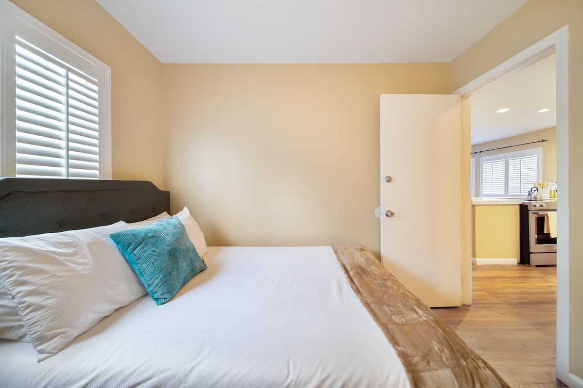 A cozy bedroom with a bed featuring a dark headboard and white bedding with a teal accent pillow, light beige walls, and white plantation shutters on the window. A doorway leads back to the kitchen area.
