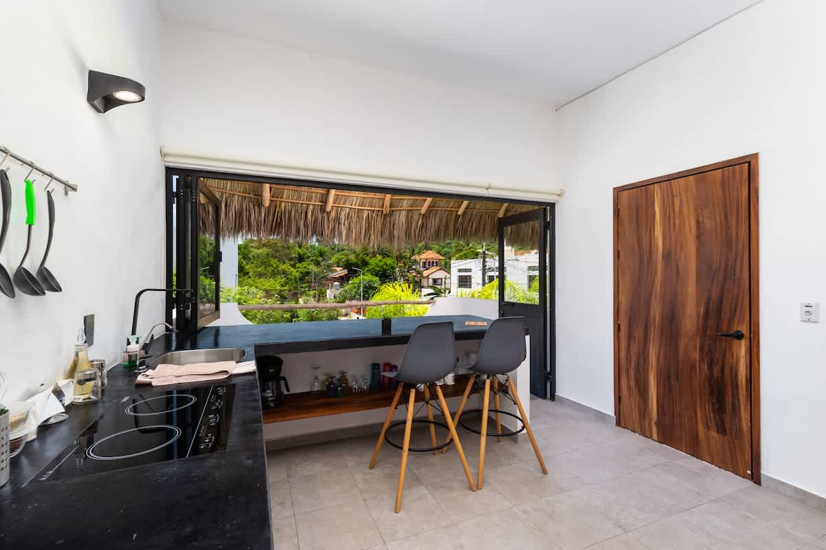 A bright kitchen features dark countertops with an integrated electric stovetop and sink, two grey bar stools, and a large wooden door, all opening to a lush outdoor view with a thatched roof, blending indoor and outdoor living within a studio loft.