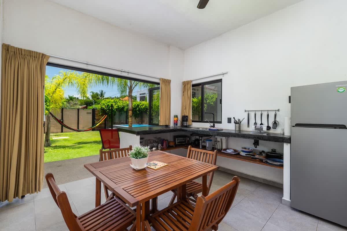 A functional kitchen and dining area with dark countertops, a stainless steel refrigerator, and a wooden dining set. Large glass doors offer a direct, inviting view of a lush backyard with a swimming pool and hammock.