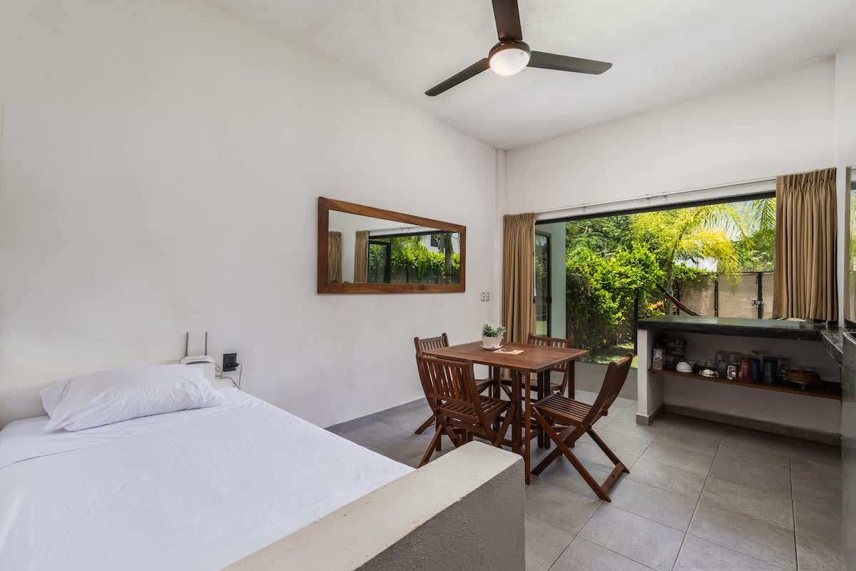 White-walled studio space featuring a bed with white linens, a wooden dining table with four chairs, and a compact kitchen counter with a large window overlooking lush greenery. A ceiling fan hangs above the dining area.