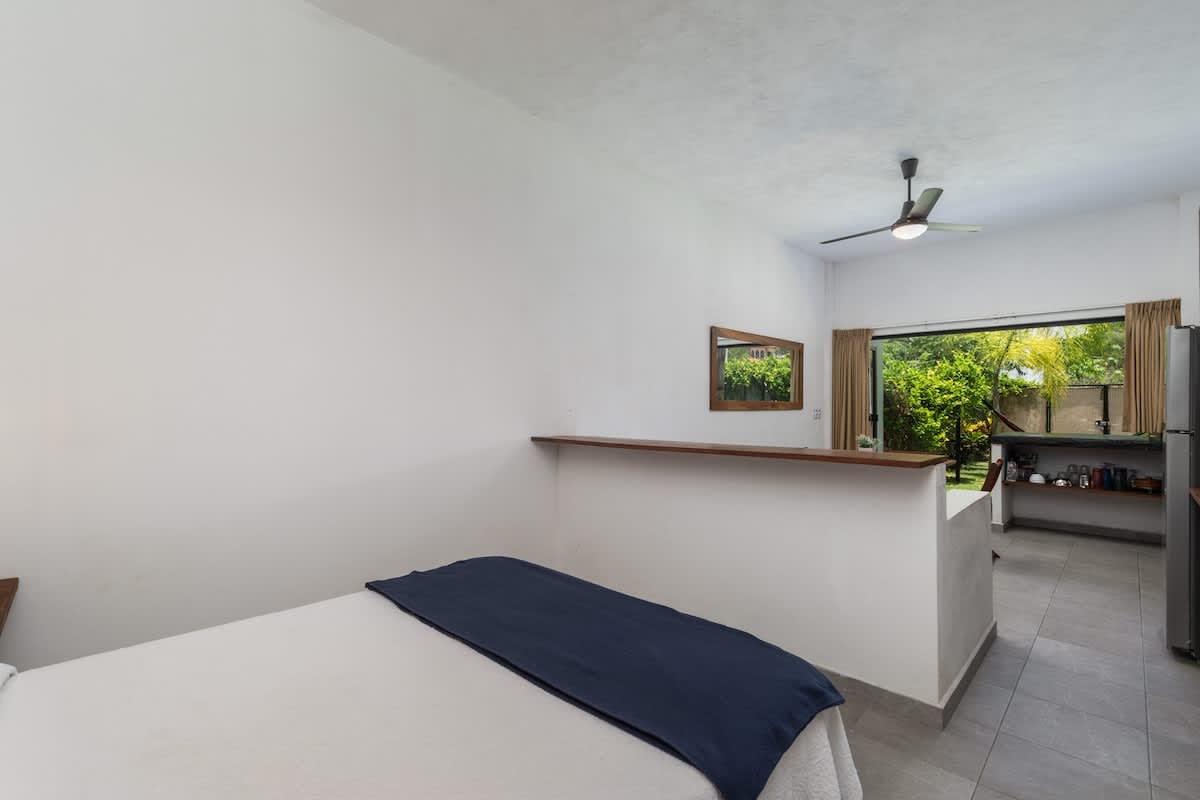 A wide view of an open-plan studio, showcasing a queen bed with white bedding and a blue throw, separated by a low wall from a kitchen and dining area with a large window overlooking greenery.