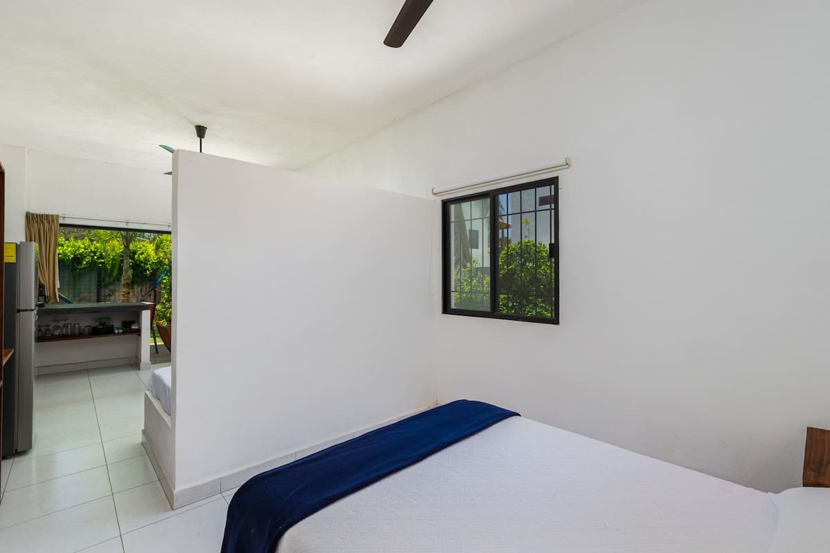 A bright, open-concept bedroom featuring a queen-sized bed with white bedding and a blue throw, separated by a low partition wall from a kitchen area. A window with black grates offers a view of greenery.