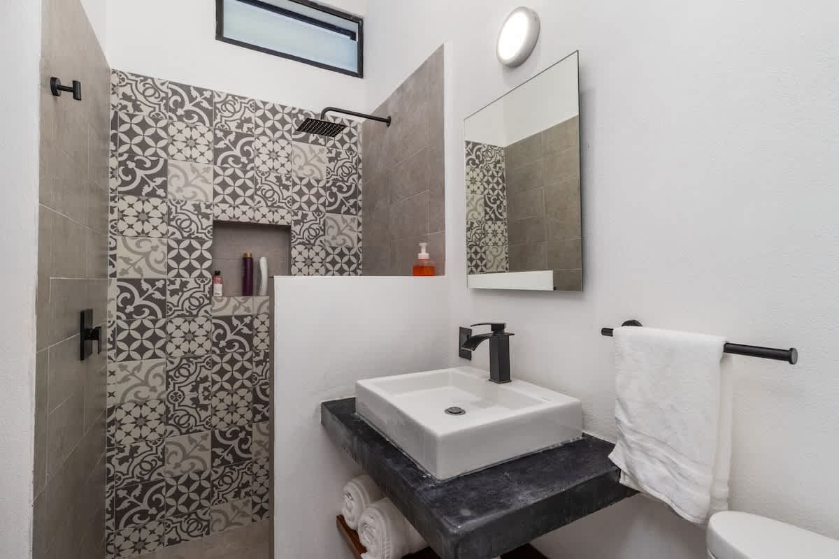 A contemporary bathroom features a walk-in shower with decorative patterned tiles and a rainfall showerhead, alongside a square vessel sink on a dark vanity with rolled white towels below, showcasing a sleek, modern design.