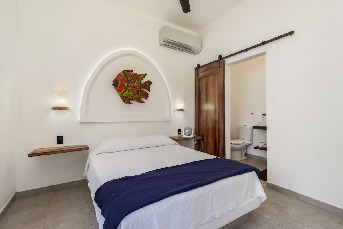 A cozy bedroom featuring a double bed with white linens and a blue throw, set against a white arched alcove with a colorful fish art piece. A dark wooden barn door leads to an en-suite bathroom.