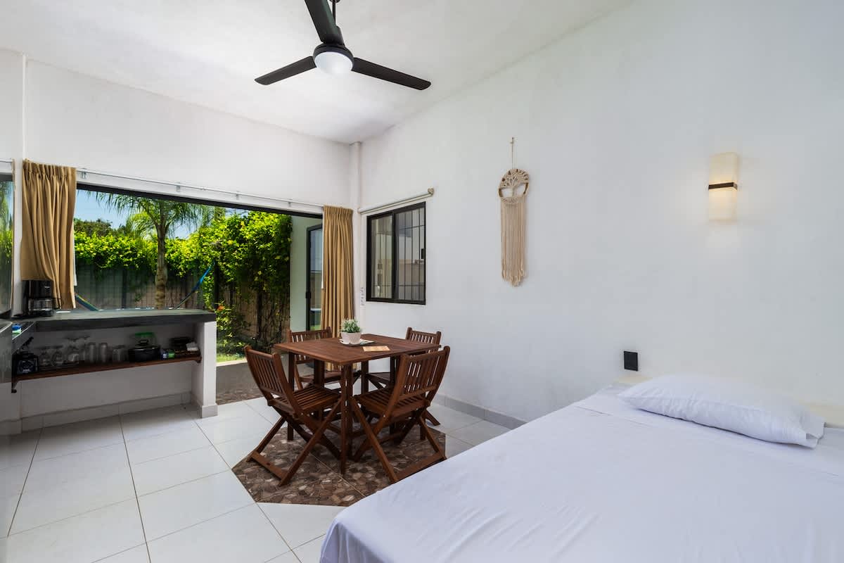 A functional kitchen and dining area with dark countertops, a stainless steel refrigerator, and a wooden dining set. Large glass doors offer a direct, inviting view of a lush backyard with a swimming pool and hammock.