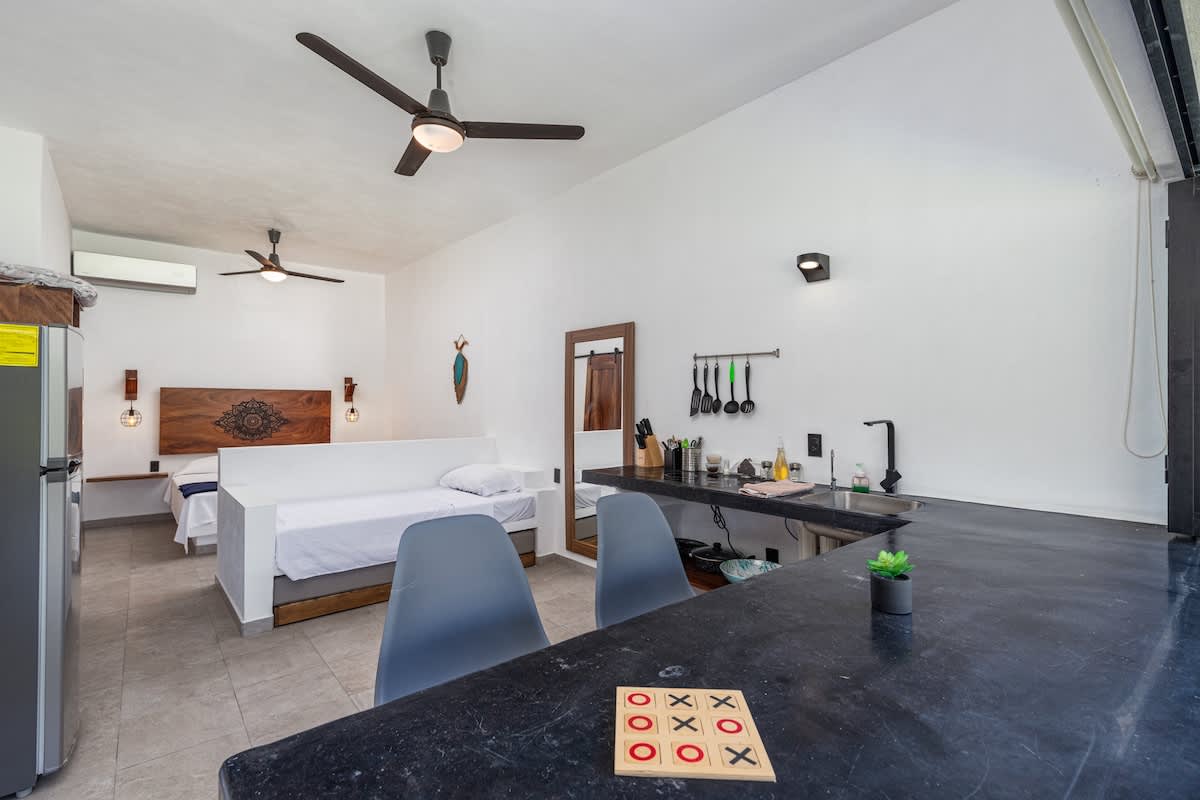 A spacious studio loft combines a modern kitchen with dark countertops and bar seating, a single bed in the foreground, and a large bed with a wooden headboard in the background, separated by a low white wall, creating an open and functional living space.