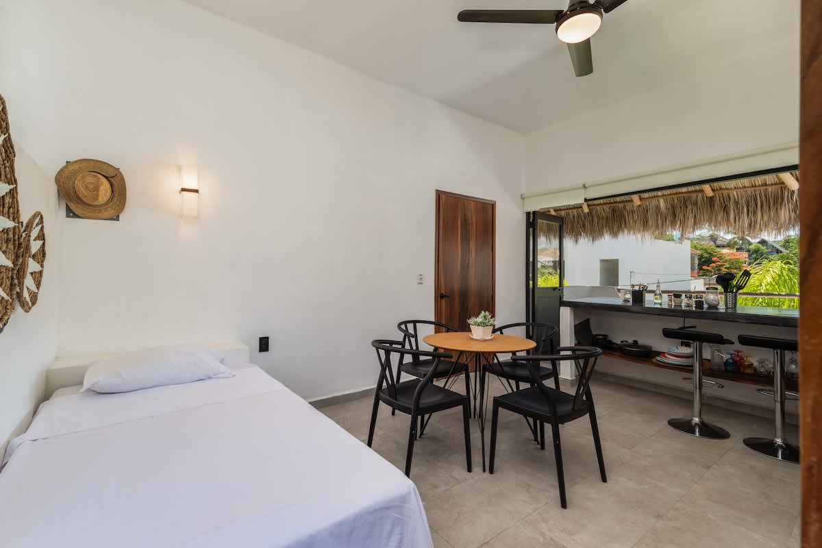 A compact studio space featuring a single bed with crisp white linens, a round wooden dining table with four black chairs, and a partial view of a kitchen counter. White walls and a ceiling fan complete the bright aesthetic.