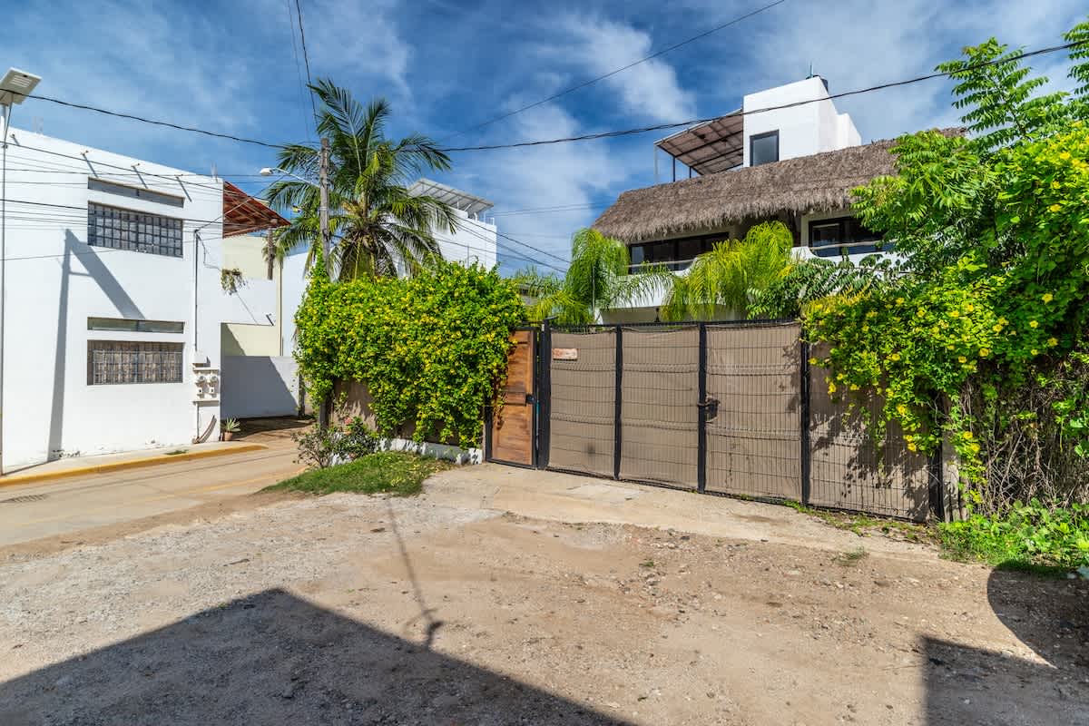 The property entrance features a large, dark metal gate with a wooden accent, surrounded by lush green foliage and a dirt road, leading to white buildings under a bright blue sky, indicating a private and tropical setting.