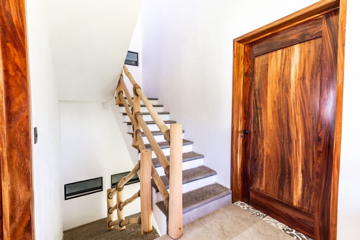 A bright interior staircase with light grey steps and a rustic bamboo railing, leading upwards. A large, richly grained wooden door is visible on the right, next to patterned floor tiles.