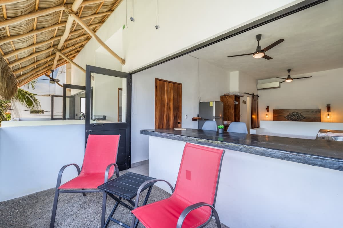 An inviting outdoor patio features two bright red chairs and a small black side table, overlooking an open-concept kitchen with a dark countertop and a thatched roof overhead, offering a relaxed tropical ambiance.