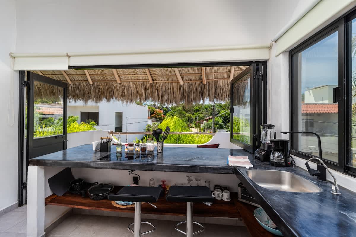 Bright and airy open-concept kitchen with dark concrete countertops, integrated cooktop, and a stainless steel sink, offering an expansive view of the tropical surroundings through large windows and an open pass-through.