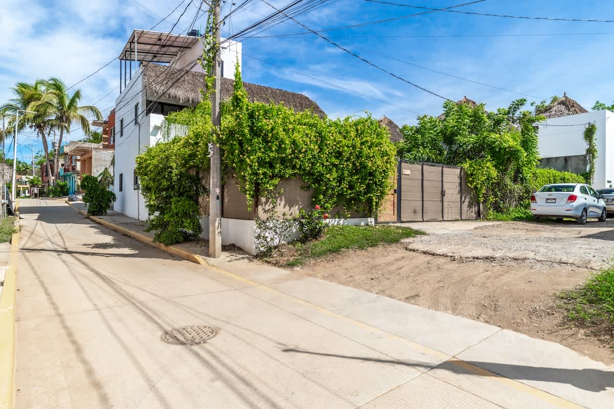 A street view showing the side of the white two-story house with a thatched roof, surrounded by lush green foliage. A paved road with a sidewalk runs in front, and power lines are visible overhead against a blue sky.