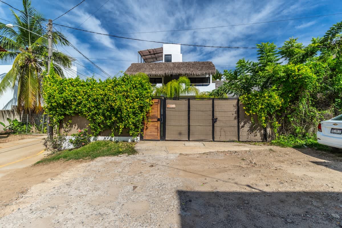 The front of a white two-story house with a thatched roof, mostly hidden behind a concrete wall covered in dense green climbing plants and a dark metal gate. A wooden pedestrian gate is on the left, and a dirt and gravel driveway is in the foreground.