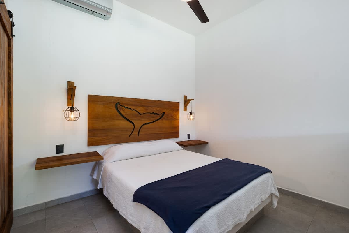 A stylish bedroom featuring a queen-sized bed with a unique wooden headboard carved with a whale tail design, complemented by floating wooden shelves and industrial-style wall sconces.
