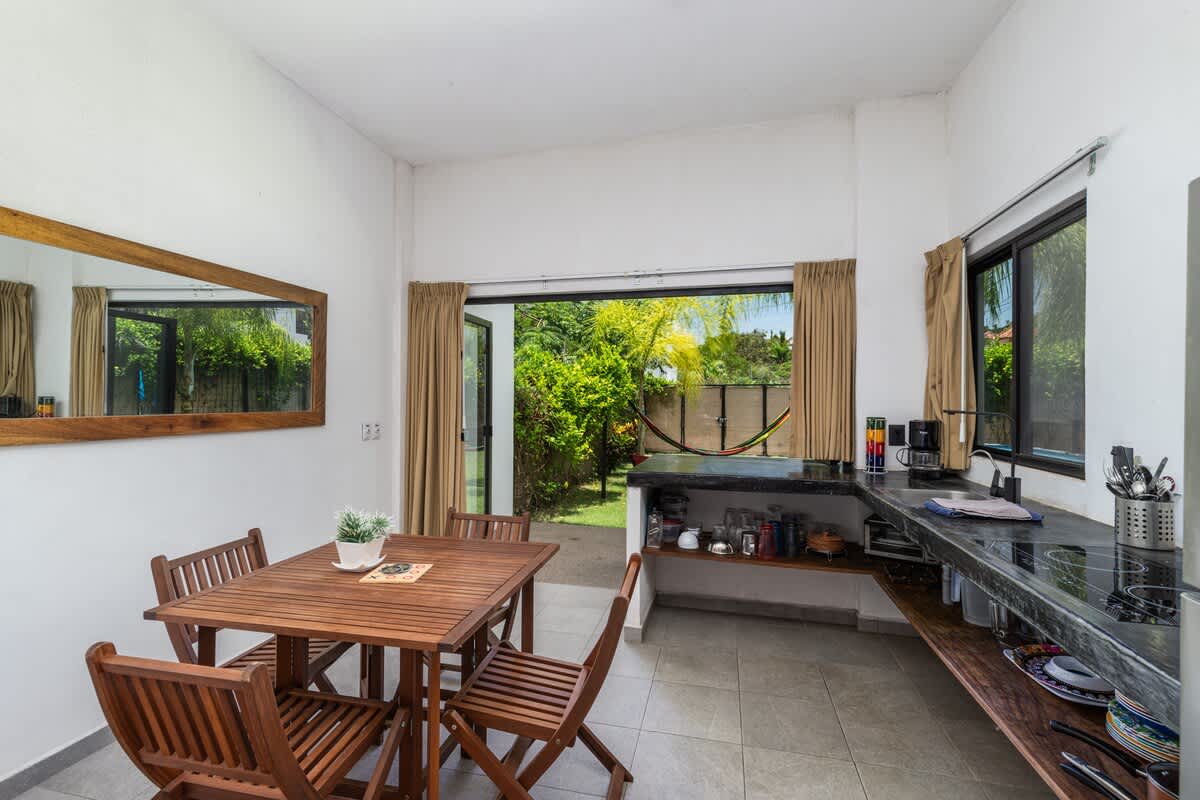 A dining space featuring a wooden table and four chairs, positioned in an open area adjacent to a kitchen with dark countertops. Wide glass doors provide an expansive view of the vibrant green outdoor garden.