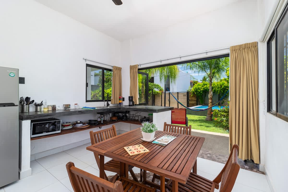 A dining space featuring a wooden table and four chairs, positioned in an open area adjacent to a kitchen with dark countertops. Wide glass doors provide an expansive view of the vibrant green outdoor garden.