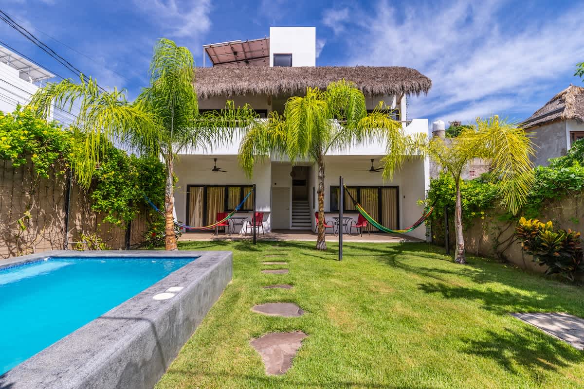 The backyard of the property, featuring a rectangular swimming pool with blue tiles and a concrete surround, a lush green lawn, and several palm trees. The white two-story house with a thatched roof and balconies is visible in the background.