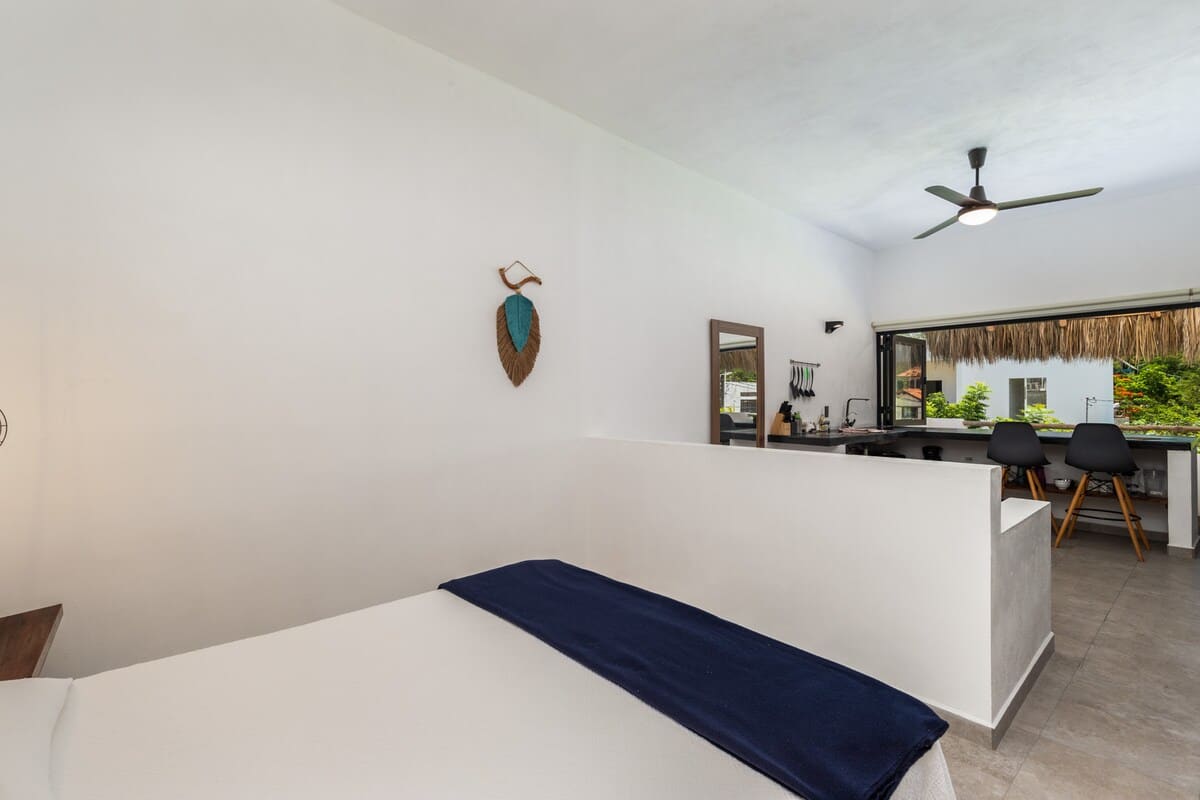 This spacious bedroom, featuring a white bed with a blue throw blanket, is partially separated by a low white wall from an adjacent open area, offering an expansive feel with an outdoor view through large windows.
