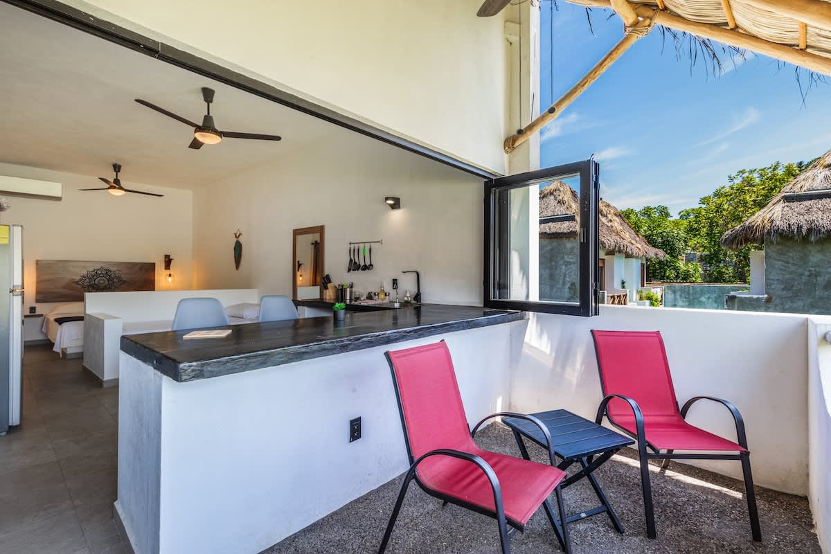 An inviting outdoor balcony with two red chairs and a small table, connected to an indoor kitchen bar area with stools. The space is open, featuring a thatched roof and views of the surrounding property.