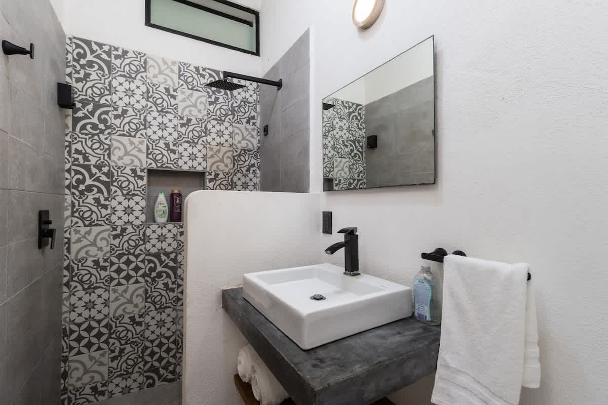 Modern bathroom with a square white vessel sink on a concrete vanity, a large mirror, and a walk-in shower featuring decorative patterned tiles and a rainfall showerhead. Black fixtures provide a contemporary contrast.