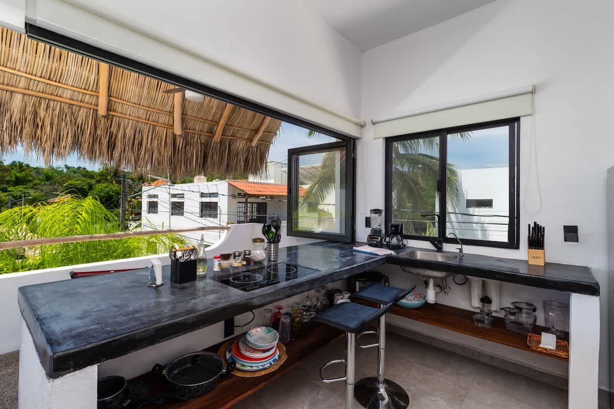 Modern open-air kitchen featuring dark concrete countertops, an induction cooktop, and a stainless steel sink, with large windows and bi-fold doors opening to a tropical view. Bar stools are tucked under the counter.