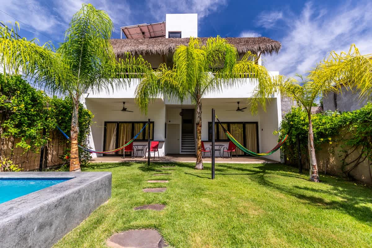 A modern two-story white building with a thatched roof on the upper level, featuring multiple ground-floor patios with hammocks and red chairs. A small rectangular concrete pool is in the foreground, surrounded by a manicured green lawn and tall palm trees under a bright sky.