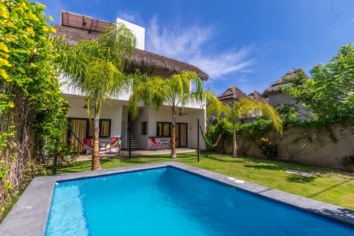 A wide shot of the backyard featuring a rectangular swimming pool with blue tiles and a concrete surround, set against a lush green lawn. The white two-story house with a thatched roof is visible in the background, with palm trees and hammocks.
