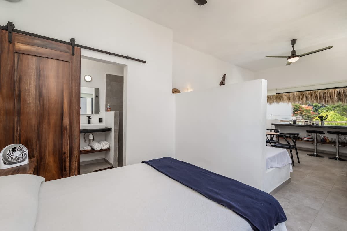 A bright bedroom with a double bed, separated by a partial white wall from the main living area. A dark wooden barn door slides open to reveal an en-suite bathroom with a sink.