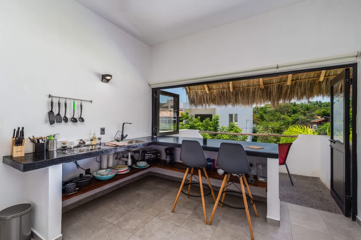 A modern kitchen with dark countertops and a stainless steel sink is open to the outdoors, featuring two grey bar stools at a counter overlooking lush greenery and a thatched roof, creating an airy, integrated space within a studio loft.