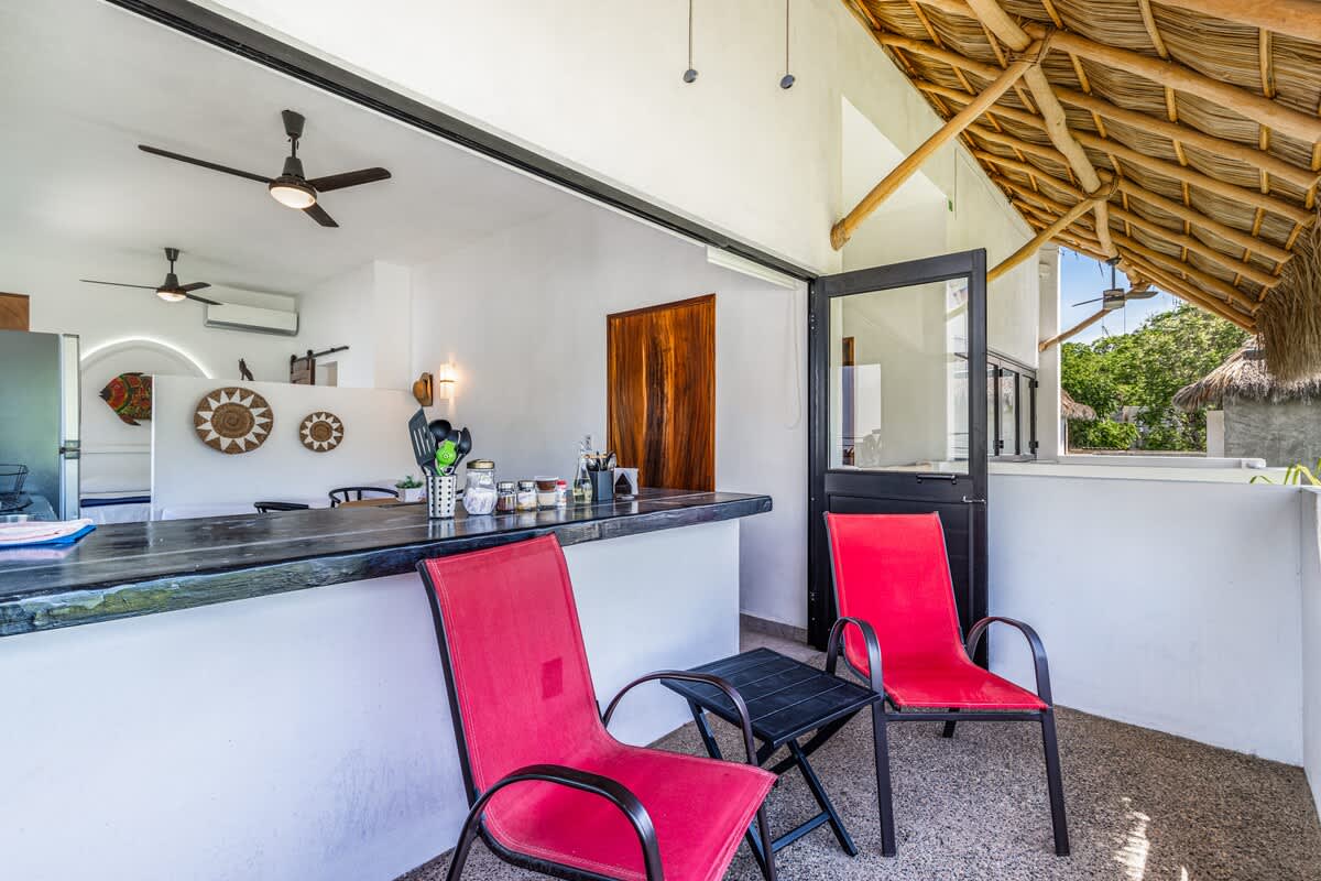 A bright outdoor balcony area with a rustic thatched roof, featuring two red metal chairs and a small black table, overlooking lush greenery. It connects to an indoor kitchen/bar space through a wide opening.